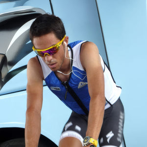 Close-up of a male cyclist in yellow Giro sunglasses and white earbuds, wearing a blue and white vest, leaning forward against a light blue backdrop.
