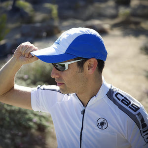 Man in a white and blue cycling cap, white sunglasses, and white Castelli jersey with "CGS" text, adjusting his hat outdoors.