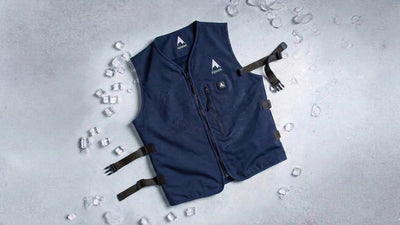 A dark blue cooling vest with a mountain peak logo and front zipper lies on a grey textured surface, surrounded by ice cubes and water droplets.