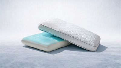 A white rectangular pillow with a geometric pattern and dark piping is angled over a flat, light blue pillow with white dots, on a cool-toned surface.