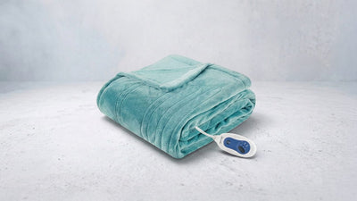 A teal plush heated blanket, neatly folded, rests on a light grey surface with its white and blue control unit visible to the right.