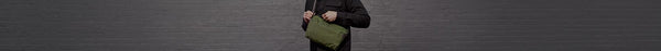 A person in a black top holds an olive green fabric sling bag with both hands against a dark grey brick wall.