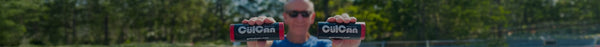 Bald man in sunglasses holds two black and red CülCan Black Ice cylindrical coolers with 