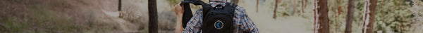 Rear view of a person in a blue and brown plaid shirt and black backpack with a 'VAPOR COOLING SYSTEM' logo, hiking on a dirt path in a blurred forest.
