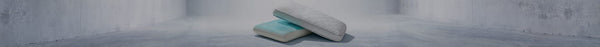 Two stacked rectangular pillows, the bottom one white with a light blue top, the top one light gray with a chevron pattern, on a grey floor.
