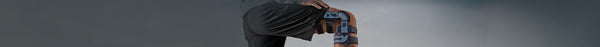 A person's bent leg in black shorts wears a black and grey knee brace, with a hand resting on their upper thigh against a grey background.