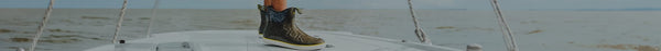 Olive green HUK boot with a yellow stripe on a white boat deck, ropes visible, calm water extends to a hazy horizon under a pale sky.