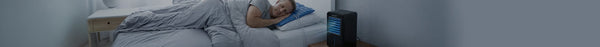 A man sleeps soundly in a dim bedroom, covered by a grey duvet, with a blue cooling pillow and a black portable cooler emitting blue light nearby.