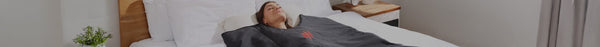 A woman with closed eyes lies peacefully on her back in a bed with white sheets, covered by a dark blanket featuring a red flame logo.