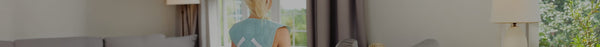Blonde person, seen from behind, wearing a light blue top with a white logo, stands in a sunlit room by a window overlooking lush greenery.