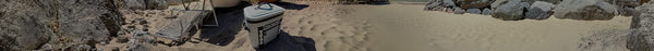 Panoramic view of a sunny rocky beach with two white lounge chairs, a grey cooler, and rippled sand, flanked by large boulders.