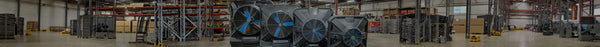 Panoramic view of a large, well-lit warehouse with industrial shelving racks, machinery, and stacks of boxes. In the foreground, several grey Portacool evaporative coolers with blue fans are displayed.