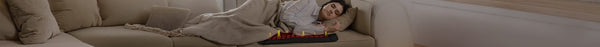 A woman sleeps peacefully on a beige sofa under a blanket, using a red and black heating device with glowing yellow indicators.