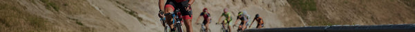 A panoramic view of several cyclists racing on a dusty path. A focused cyclist in red and black leads on the left, with blurry riders behind on dry, sunny hills.