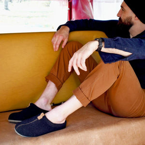 Men's algae-infused wool slippers by Acorn in black and dark gray with brown labels, worn by a man on a brown couch.