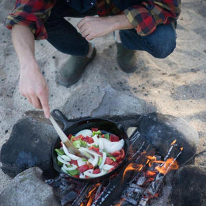 Dark green men's moccasins by Acorn, worn by a person cooking over a campfire on sand.