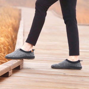 Grey knit women's moccasin slippers by Acorn, with black trim and orange heel pull, worn on a boardwalk.
