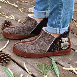 Brown felted bootie slippers by Acorn, featuring an embroidered owl and white zig-zag stitching, worn on a wooden deck.