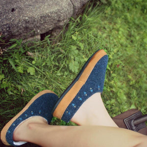 Women's blue felt hoodback slippers by Acorn, with tan trim and embroidered geometric triangles in blue and gold, worn on feet outdoors on grass.