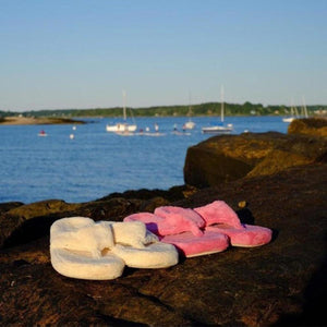 Pink fluffy Women's Spa Thong Slippers by Acorn, displayed with white slippers on dark rocks by a calm blue ocean.