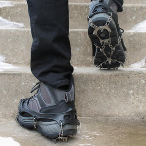 Black metal ice cleats by Action Traction, worn on hiking shoes ascending snowy concrete steps.