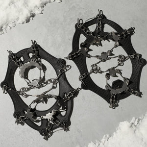 Two black rubber ice cleats by Action Traction, featuring silver metal chains and spiked plates, on a gray surface with snow.