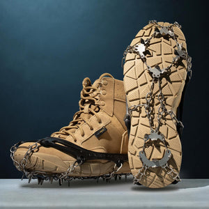 Silver metal ice cleats with black rubber straps and spikes by Action Traction, fitted onto tan tactical boots, one sole displaying rugged treads.