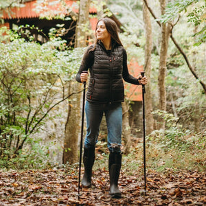 Black insulated puffer vest by ActionHeat with orange zipper pulls, worn by a woman holding hiking poles in a forest.