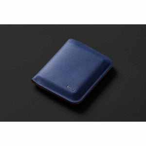 Blue smooth material note sleeve by Bellroy, closed, with a subtle debossed logo and red trim along the bottom edge, on a dark background.