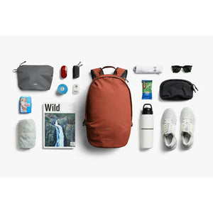 Burnt orange lightweight daypack by Bellroy, shown flat lay with various travel items beside it on a white surface.