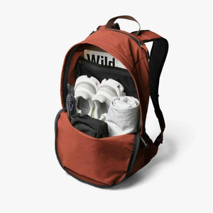 Burnt orange lightweight daypack by Bellroy, shown open with white sneakers, a rolled cloth, a small black pouch, and a 'Wild' magazine inside.