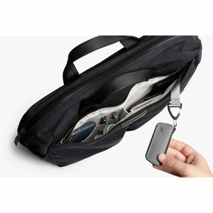 Black fabric work bag by Bellroy, open showing its light interior with items and a hand holding a key case.