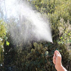 Black personal mister by ExtremeMIST spraying a fine mist, held by a hand against green bushes.