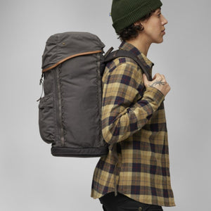 Dark olive robust backpack by FjallRaven with brown leather trim and front lacing, worn by a person in a plaid shirt and beanie, side view.