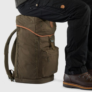 Dark olive Singi Stubben robust backpack by FjallRaven, with a person sitting on its padded top, wearing black pants and brown hiking boots.