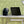 Black personal air circulator by Vornado with silver spiral fan blades on a white desk, near a laptop, against a patterned green wall.