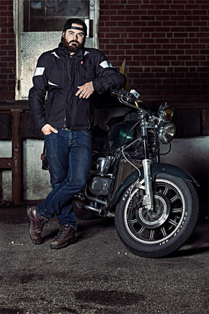 Black motorcycle heated jacket by Gerbing with white reflective strips, worn by a man leaning on a motorcycle.