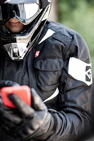 Black Motorcycle Heated Jacket by Gerbing, worn by a rider in a helmet and gloves, holding a red phone, with white accents and logos.