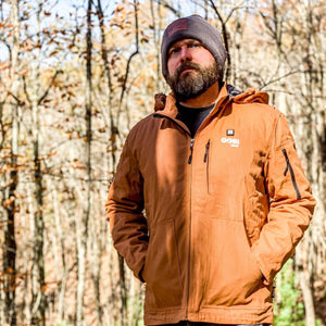 Men's rust brown heated workwear jacket by Gobi Heat, with a hood and multiple zippered pockets, worn by a bearded man in a forest.