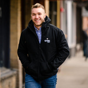 Black men's heated hoodie by Gobi Heat with a white logo, worn by a smiling man over a plaid shirt and jeans.