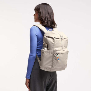 Light taupe Flap Pack Bag by Got Bag, worn on a woman's back, with a water bottle in the side pocket and colorful zipper pull.