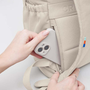 Beige flap pack bag by Got Bag, with a hand inserting a light grey phone into its zippered side pocket and a colorful tag.