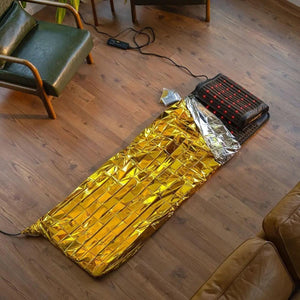 A black rectangular heated mat with red lights by HealthyLine, partially covered by a gold emergency blanket on a wooden floor.