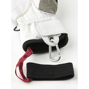 White leather glove by Hestra, featuring grey fabric, a red strap, and a silver carabiner, all showing Hestra branding.