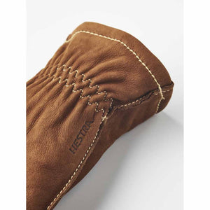 Brown nubuck leather gloves by Hestra, featuring prominent white zigzag stitching, a debossed Hestra logo, and an elasticized cuff in a cropped view.