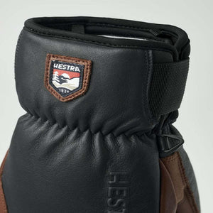 Textured black leather 3-finger glove by Hestra, featuring brown accents, a Hestra logo patch, and an adjustable wrist strap.