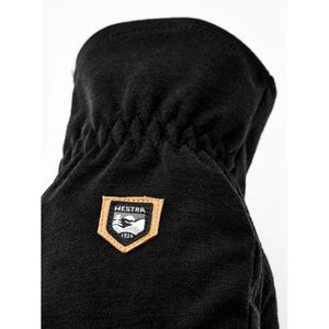 Black Merino Windwool 5-Finger Liner Glove by Hestra, featuring an embroidered patch with '1936' and textured fabric.