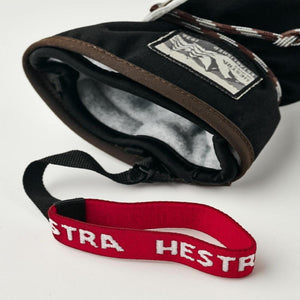 Black fabric mitten by Hestra with brown trim, white lining, a brand label, braided rope, and red logo strap.