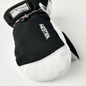 Two-tone black and white junior mittens by Hestra with a textile upper, leather lower, brown cuff, white Hestra logo, and braided cord.