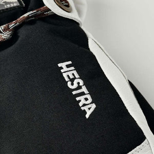 Black fabric and white leather mittens by Hestra, with an embroidered logo and a woven strap with a bronze grommet.